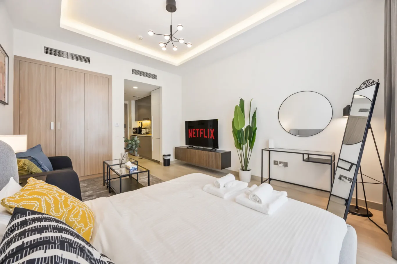Modern hotel suite: view from the bed toward a living area with a Netflix TV, a wooden wardrobe, and tall green plants.