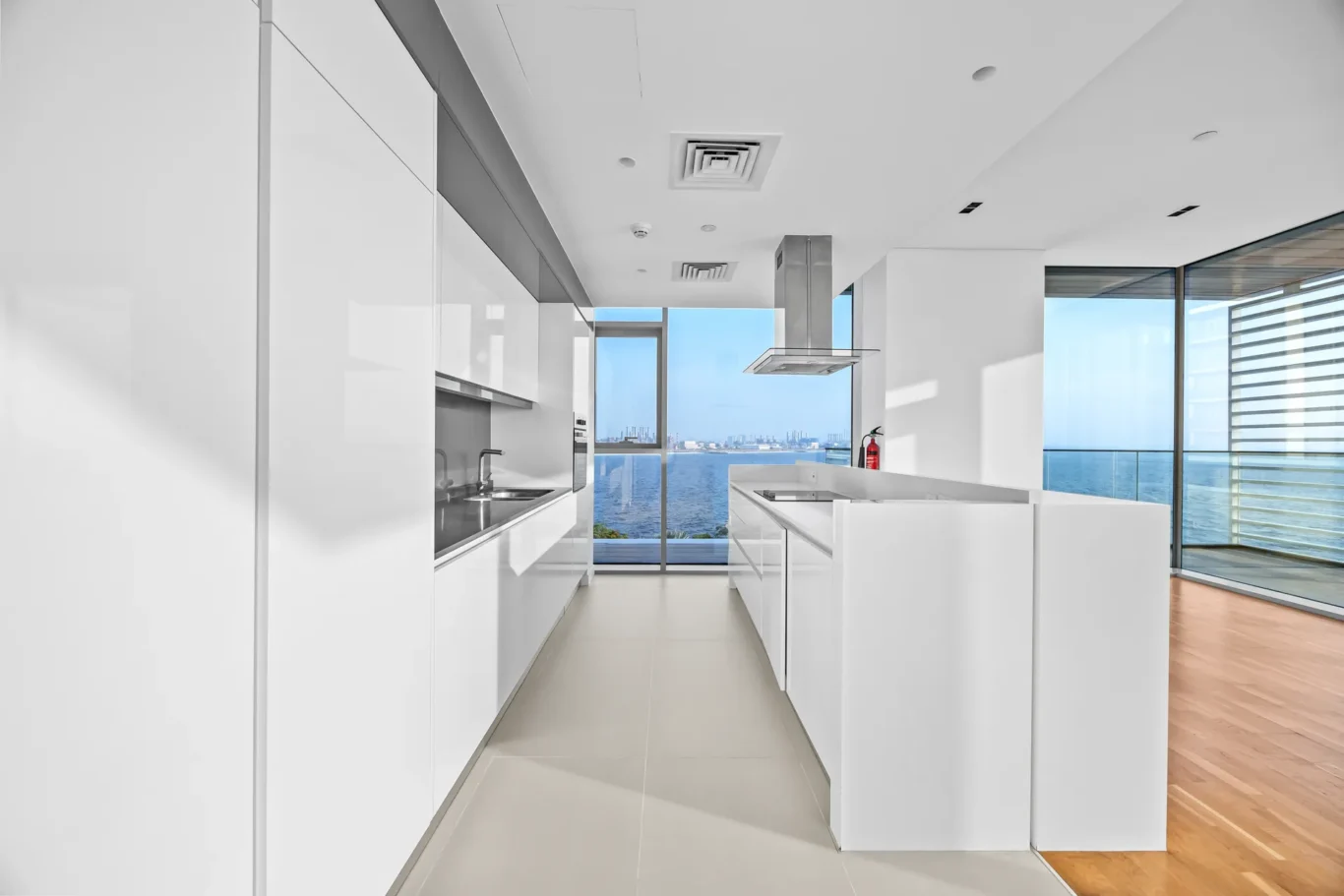Bright, modern white kitchen with a long island, sleek cabinets, and a view of the ocean through large windows.