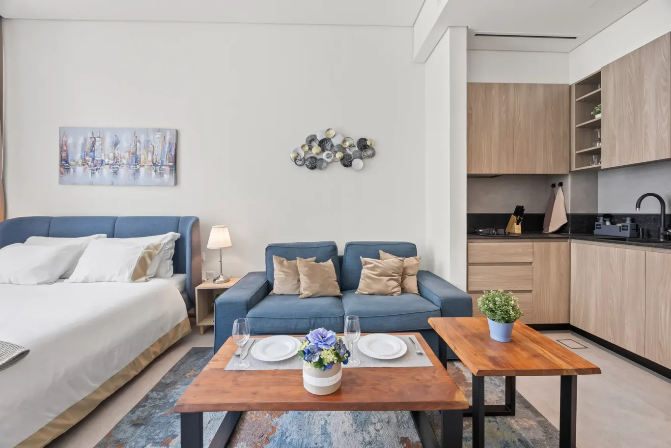 Open-plan living area with a blue upholstered bed on the left, a blue sofa with tan cushions, a wooden coffee table set for two, and a compact kitchenette with wood cabinets on the right. A small lamp and wall art decorate the wall.