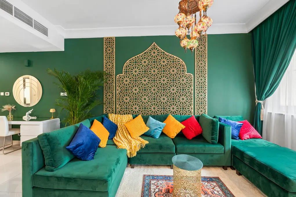 L-shaped green velvet sofa with multicolored cushions in a Moroccan-inspired living room featuring a gold lattice screen on a dark green wall.