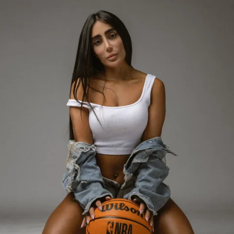 Woman kneeling on the ground holding an orange basketball between her knees, wearing a white crop top and a denim jacket draped over her shoulders against a gray studio background.