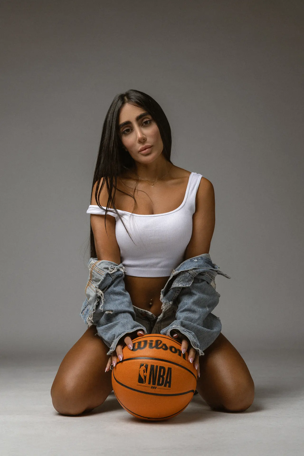 Woman kneeling on the ground holding an orange basketball between her knees, wearing a white crop top and a denim jacket draped over her shoulders against a gray studio background.