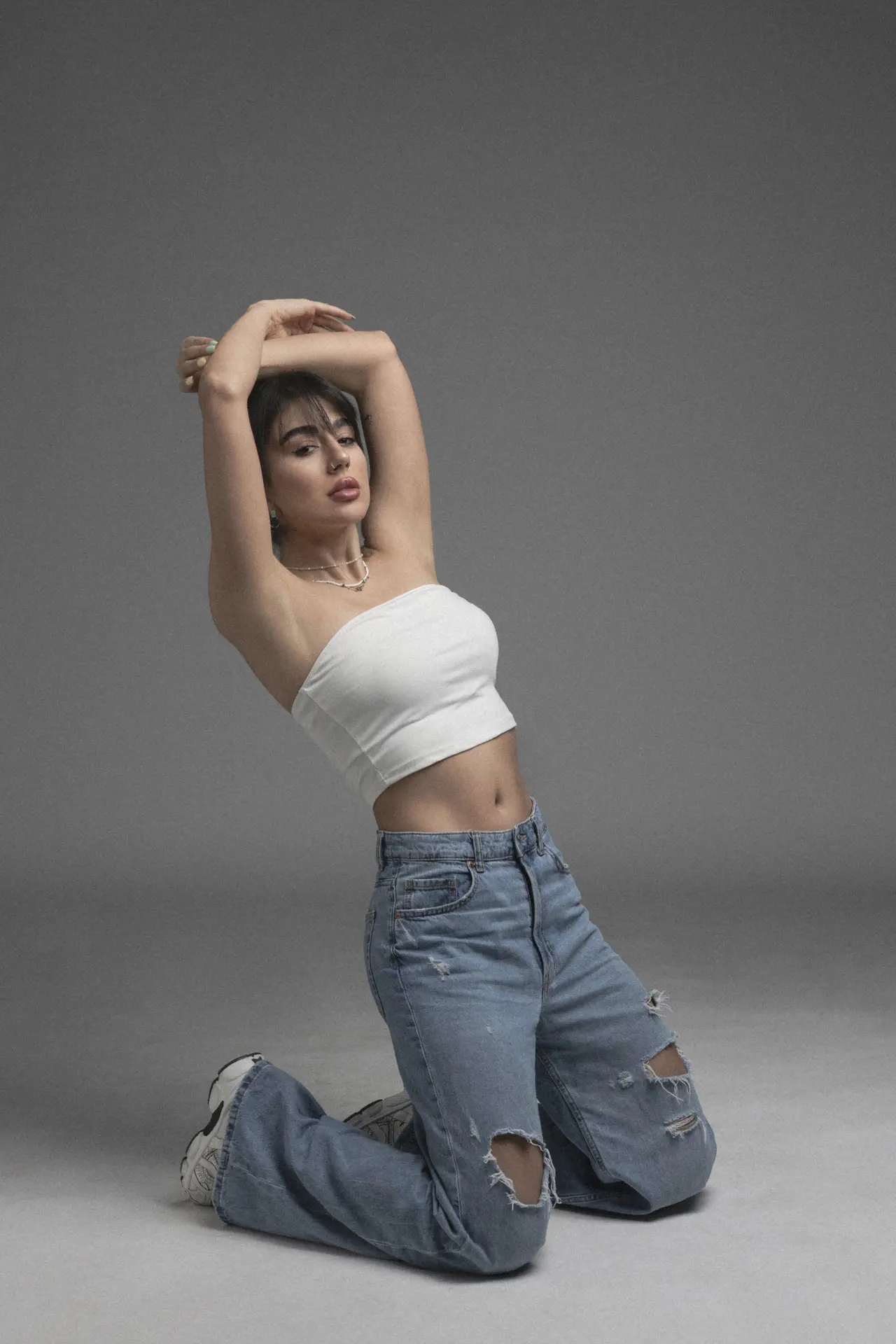 Woman in a white crop top and ripped jeans poses on a gray studio backdrop, arms raised above her head in a stretching pose.