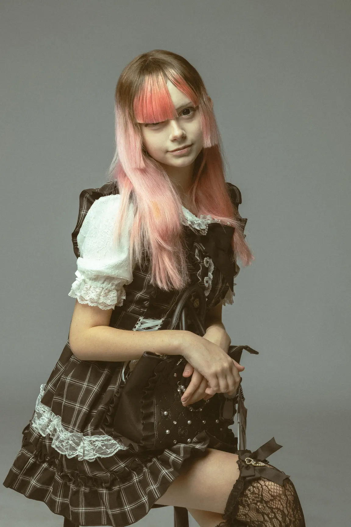 Portrait of a young woman in a black plaid Lolita-style dress with lace trim, holding a black handbag, pink dyed hair with blunt bangs, seated against a gray backdrop.