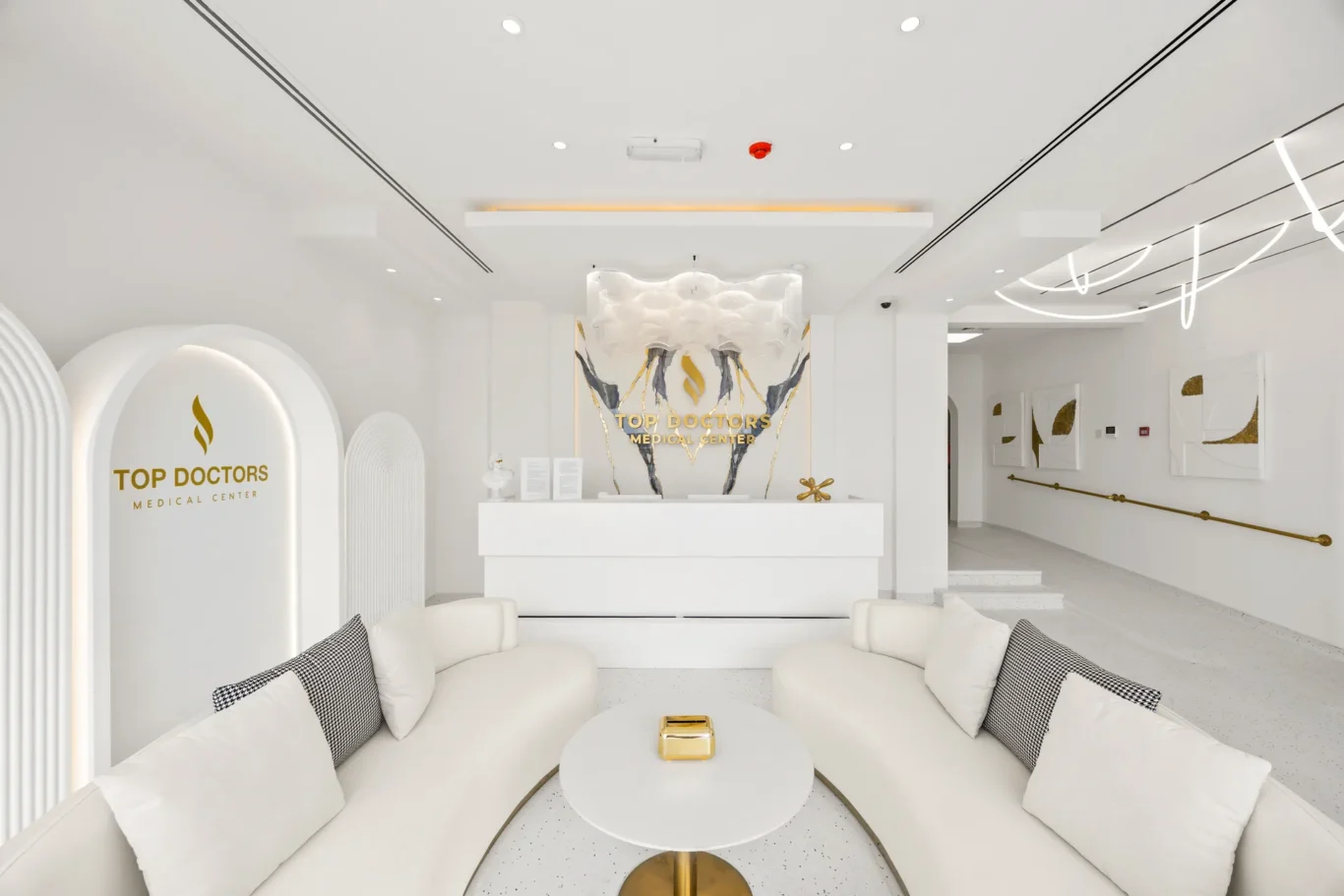 White modern medical center lobby with a curved white sofa seating area and a gold-embossed 'Top Doctors Medical Center' logo on the wall behind the reception desk.