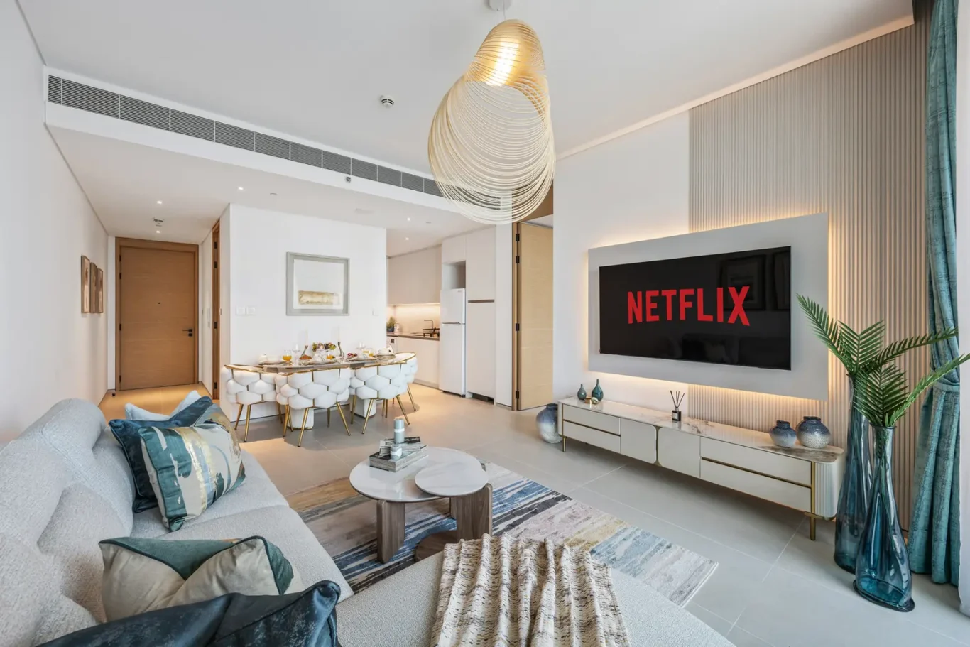 Open-plan living area with a wall-mounted Netflix screen, white media console, and teal decorative vases near a large window frameed by blue curtains.