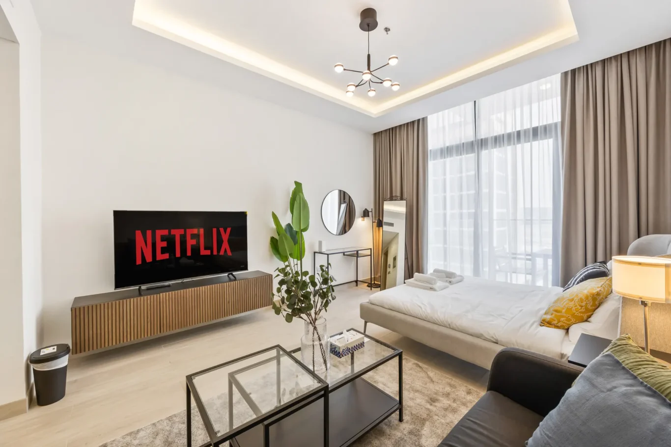 Modern living room with a Netflix TV on a wooden console, potted plants, a round mirror, and glass coffee tables by a large window with sheer curtains.