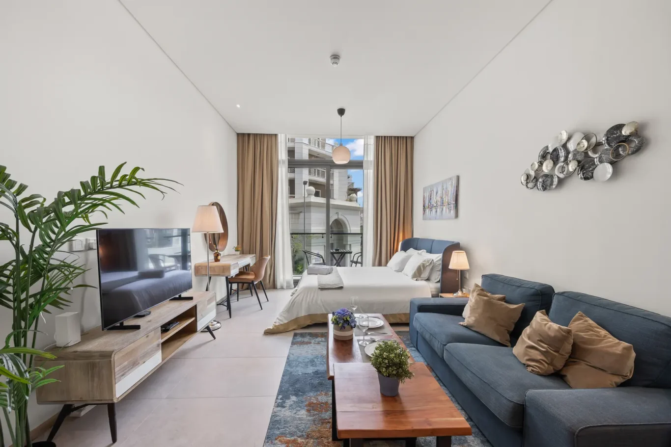 Open-plan hotel suite with blue sofa, wooden coffee table, and a bed toward the back; large balcony doors with beige curtains beyond, and a TV on a wooden console left.