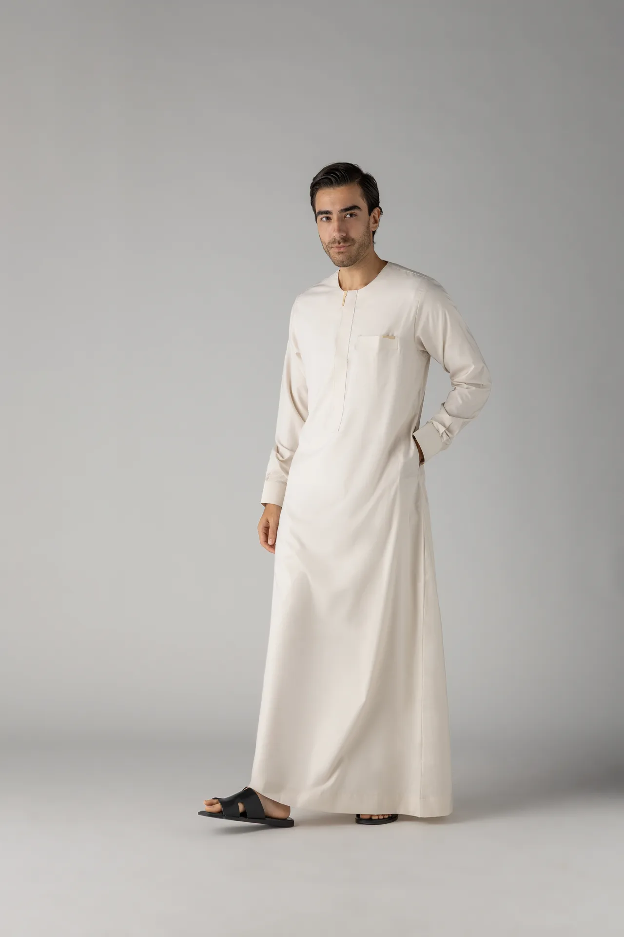 Man wearing a cream thobe with a chest pocket, standing in a studio against a gray background, black slide sandals on.