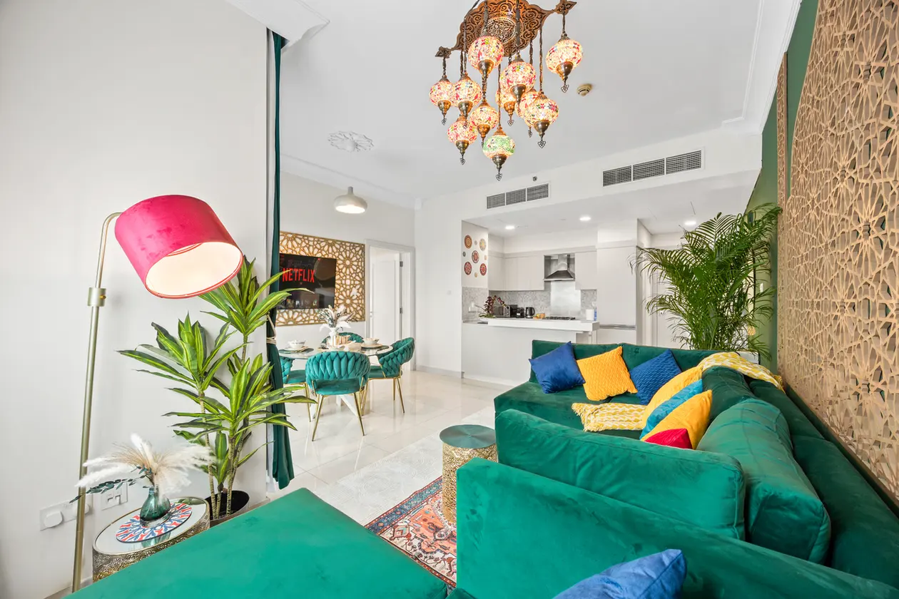 Open-concept living room with a green sofa, colorful cushions, and a patterned wall panel; dining area and kitchen in the background.