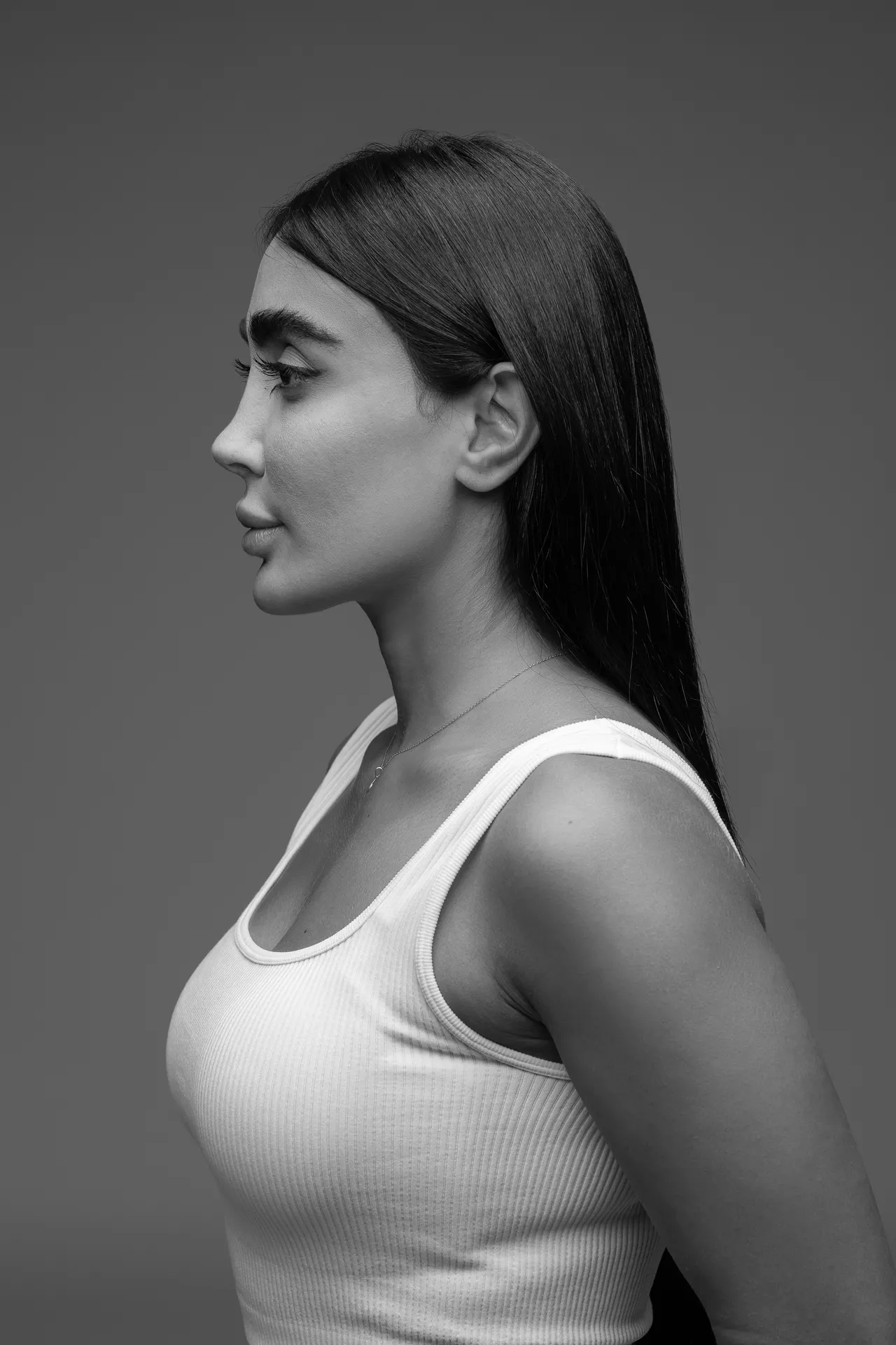 Profile view of a woman with long dark hair wearing a white ribbed tank top against a gray background.