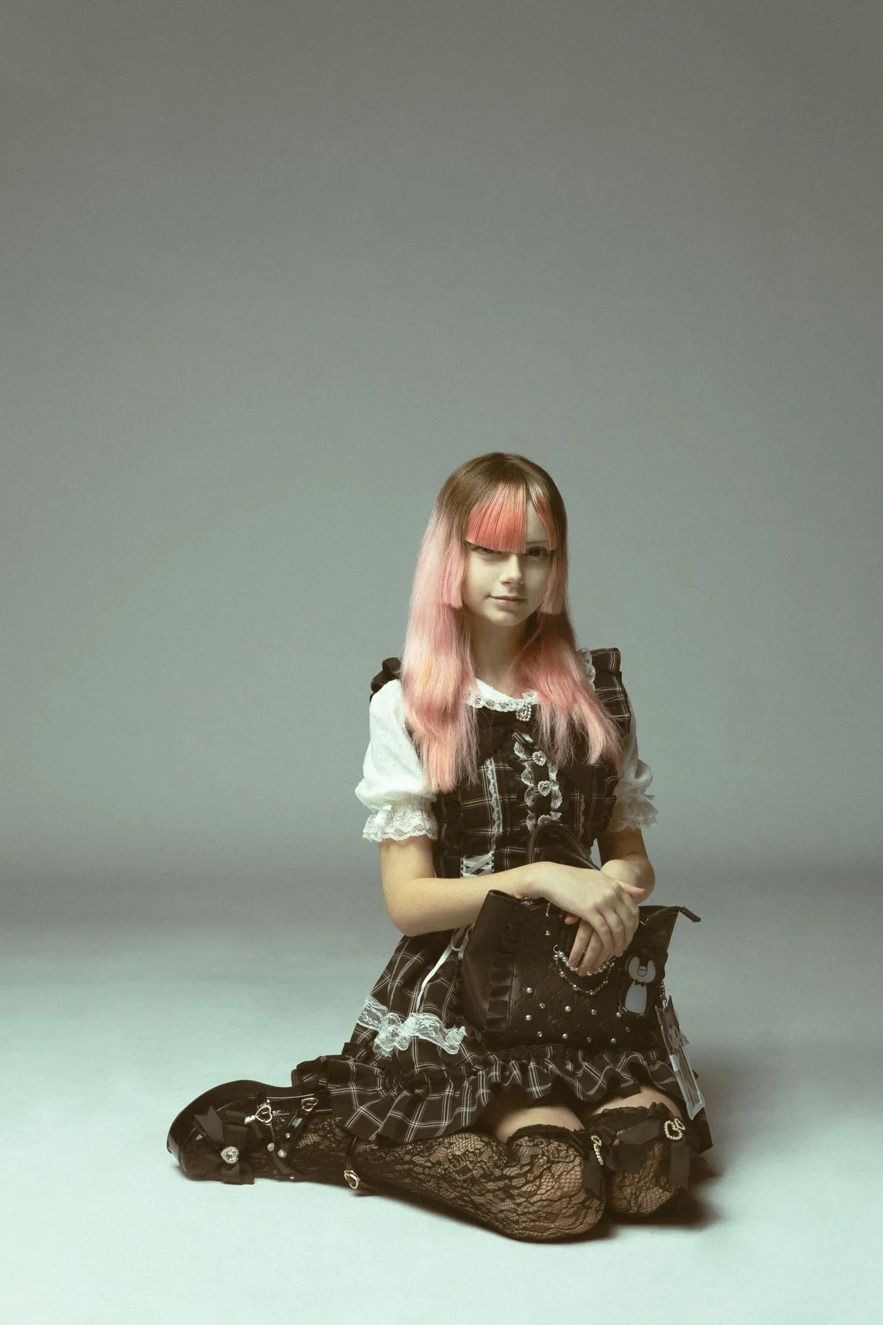 Young woman with pink hair wearing a black gothic lolita dress, sitting on the floor with a black handbag.