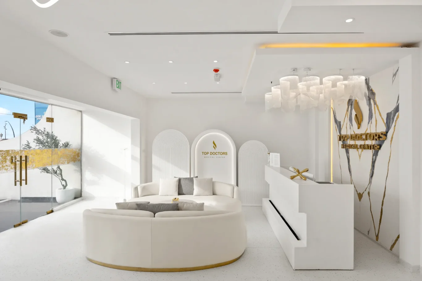 Modern white medical lobby with circular sofa, glass doors, and a reception desk; wall reads 'Top Doctors Medical Center' in gold.