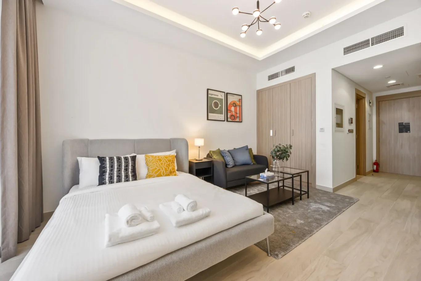 Bright hotel room with a large bed in the foreground, gray headboard, and decorative pillows; a seating area with a sofa, coffee table, and lamp nearby.