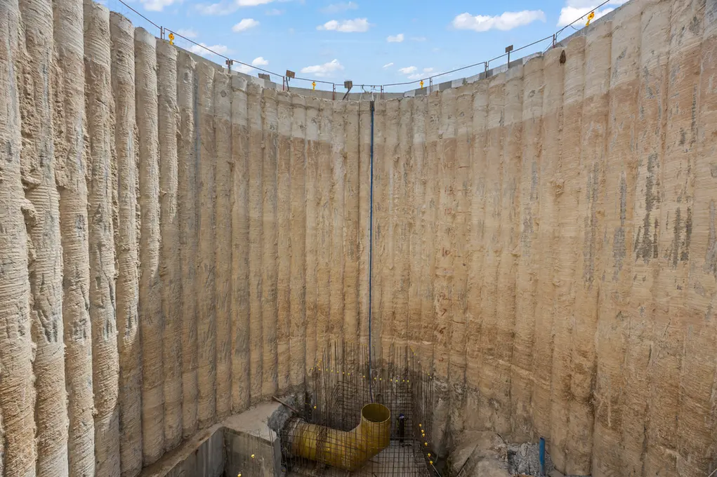 Deep circular construction shaft with tall, textured concrete walls and a blue sky above, showing a yellow drainage pipe at the bottom and exposed rebar forming a grid.
