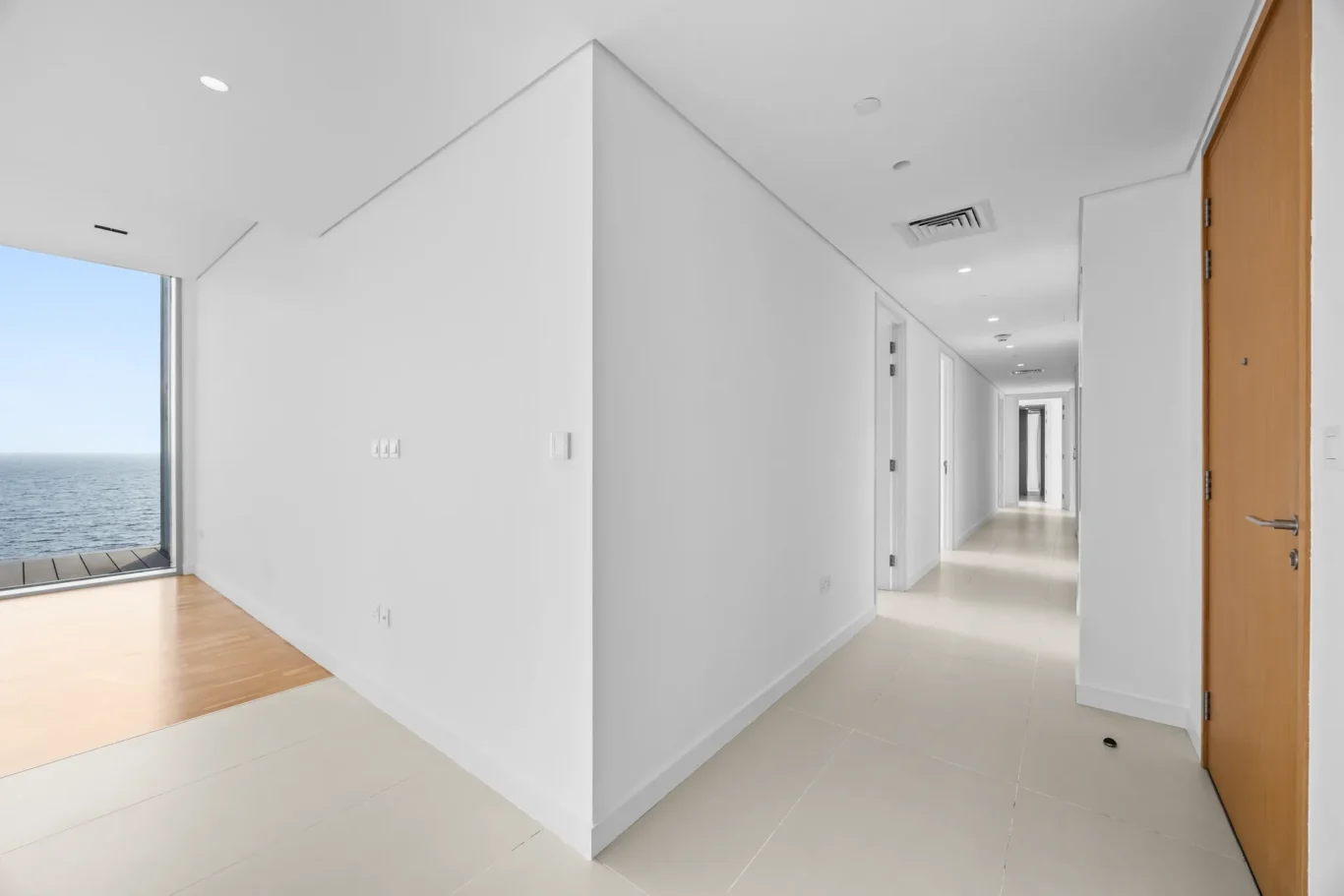 Bright white hallway with multiple doors, wooden door on the right, and a large window showing the ocean on the left.