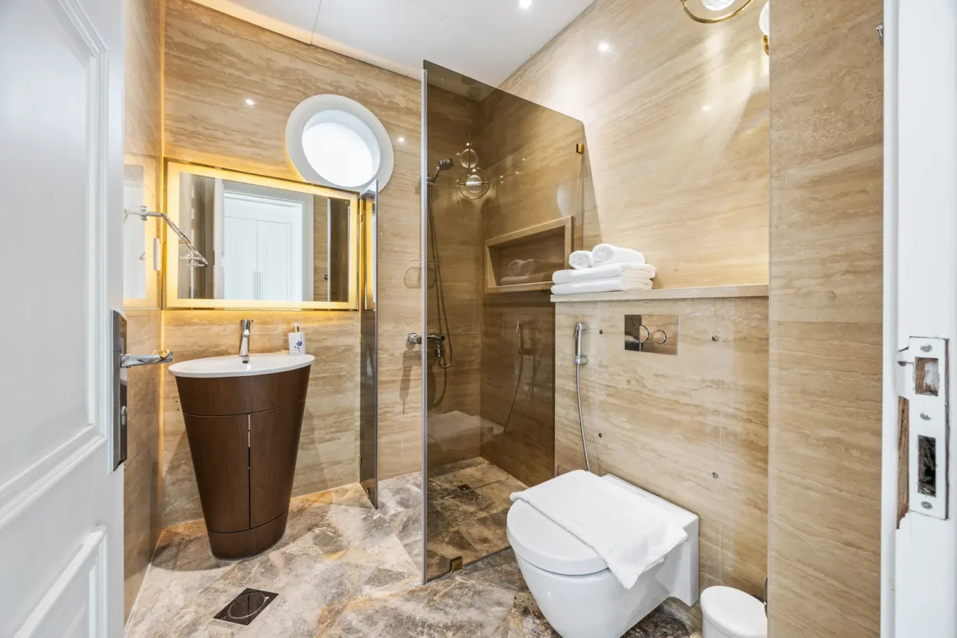 Luxurious marble bathroom with a glass-enclosed shower, round mirror, and pedestal sink under warm lighting.