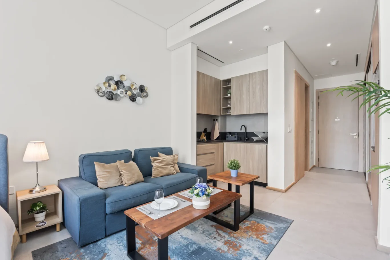 Living room with a blue sofa and beige cushions, wooden coffee table, and a kitchen opening in the background.