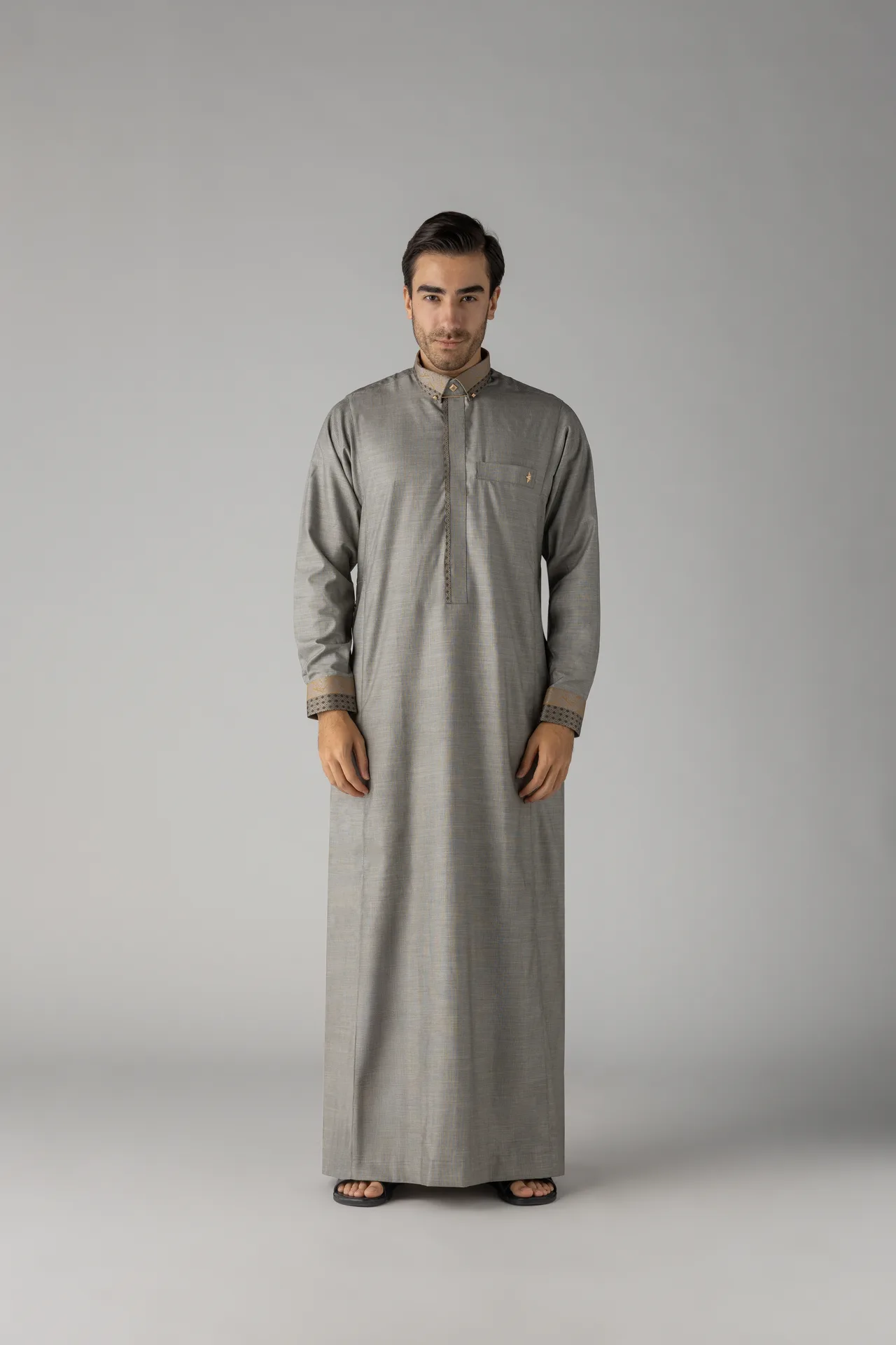 Front view of a man in a long gray kurta with beige cuffs and collar details, standing against a plain gray backdrop, wearing sandals.