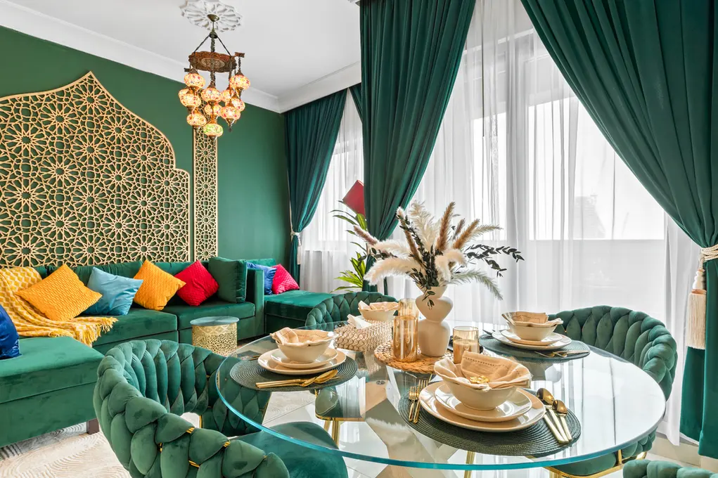 Emerald green living-dining area with tufted chairs, colorful cushions, and a round glass table set with gold utensils and bowls.