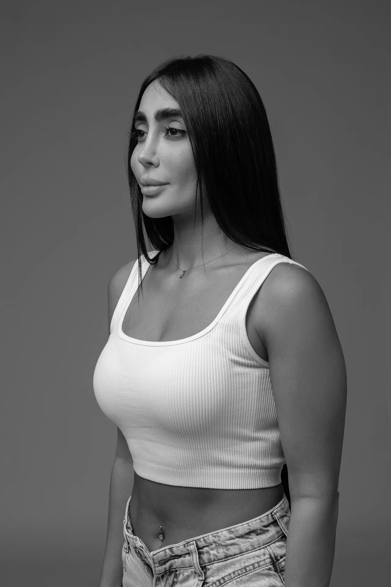 Black-and-white portrait of a woman with long dark hair, wearing a white ribbed tank top and denim jeans, facing left.