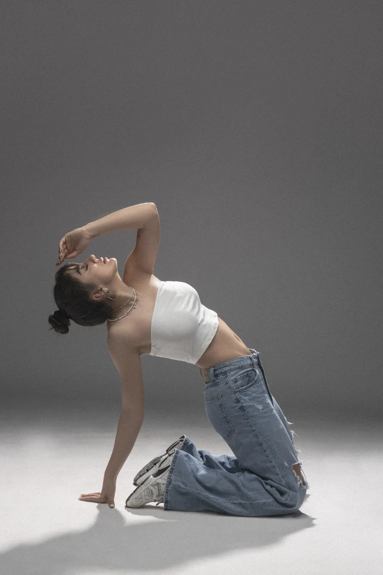 Young woman in a white crop top and jeans kneels on the floor, performing a backward backbend in a studio setting.