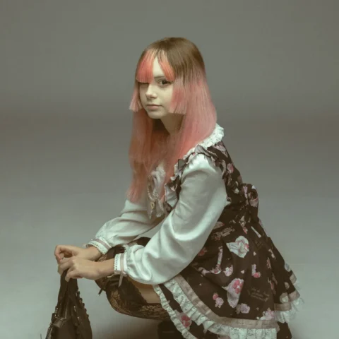 Young woman in a lace Victorian-style Lolita dress with pink gradient hair, crouching and holding a black handbag against a plain backdrop.
