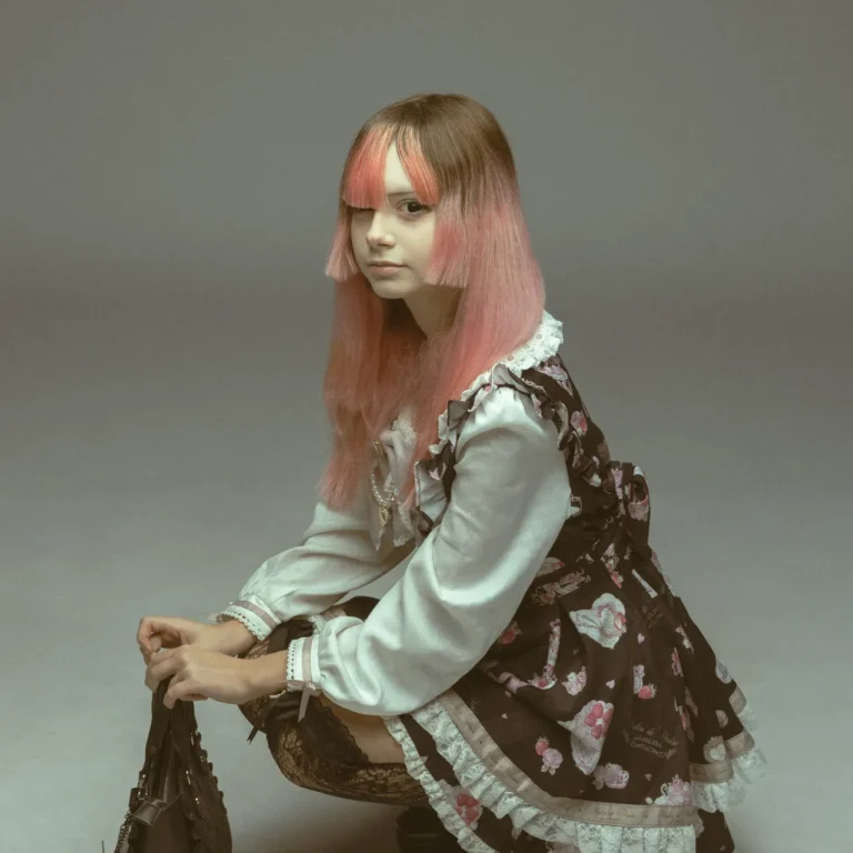 Young woman in a lace Victorian-style Lolita dress with pink gradient hair, crouching and holding a black handbag against a plain backdrop.