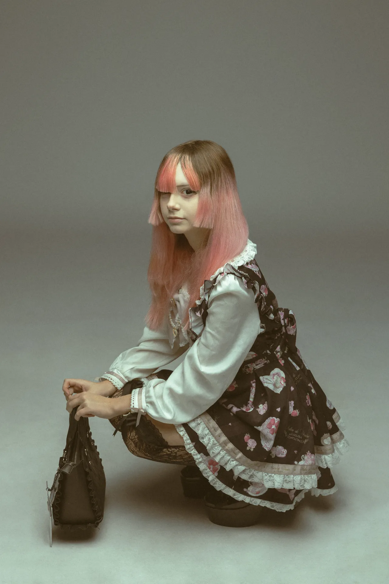 Young woman in a lace Victorian-style Lolita dress with pink gradient hair, crouching and holding a black handbag against a plain backdrop.