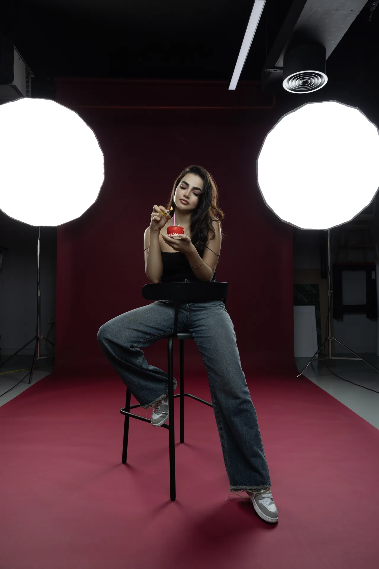 Woman seated on a tall stool in a photography studio, applying red lipstick from a small jar, with two large studio lights in the background.