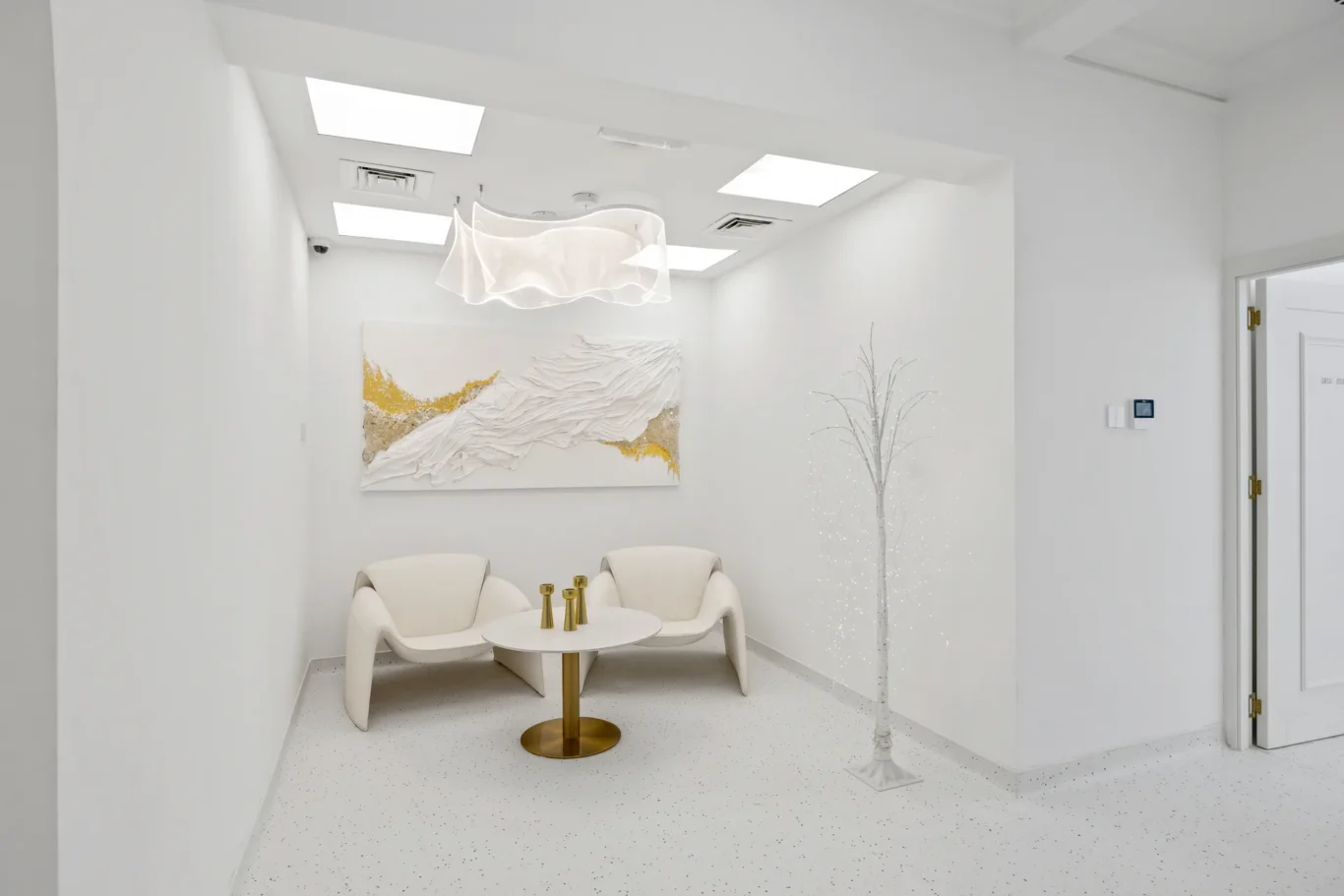 Bright white lounge with two curved chairs around a round white table with a gold base; abstract white-and-gold wall art and a lit decorative tree nearby.