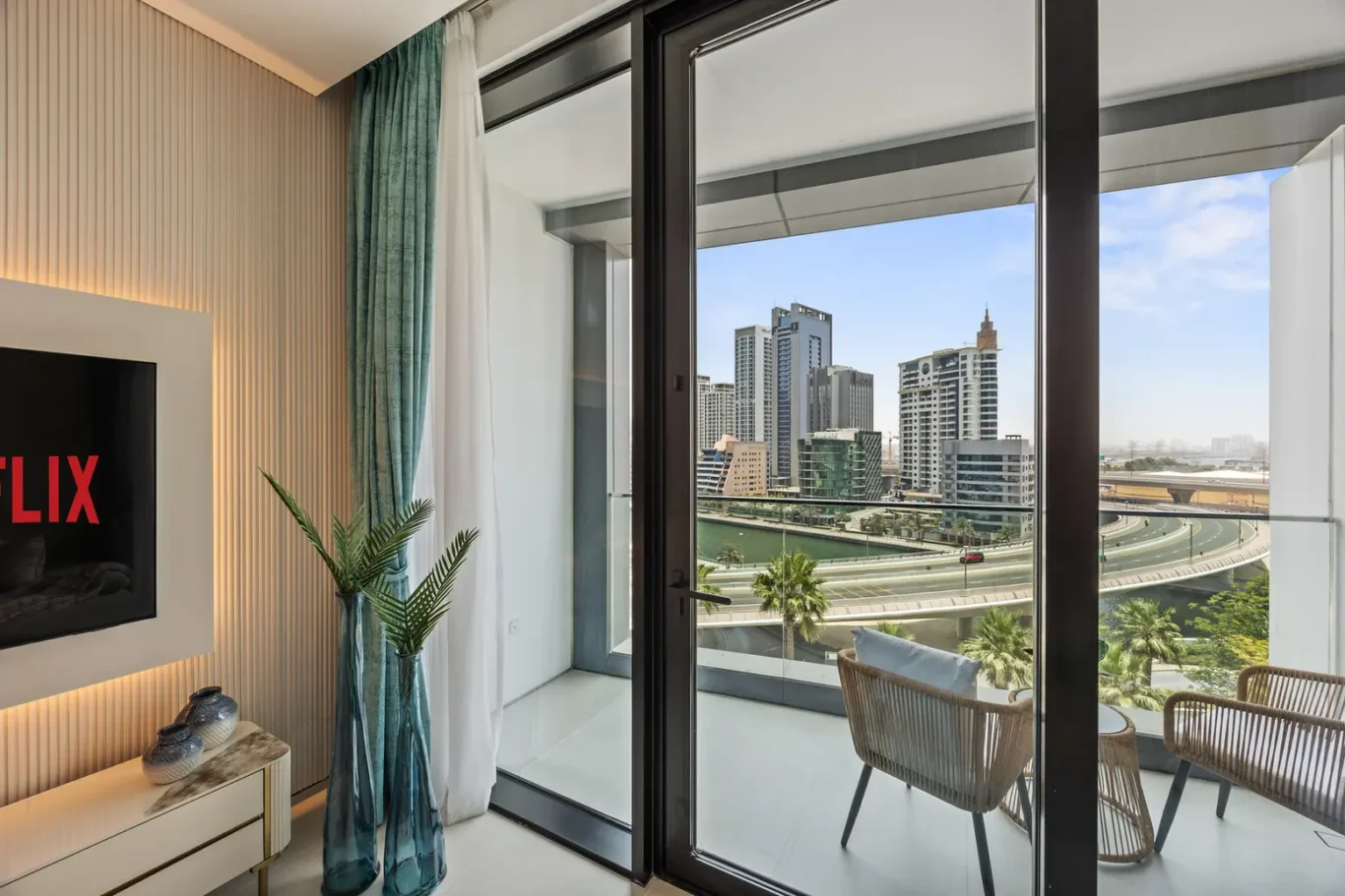 Bright living room with floor-to-ceiling glass balcony doors opening to a city view of skyscrapers and a curved highway below; wicker chairs on the balcony.