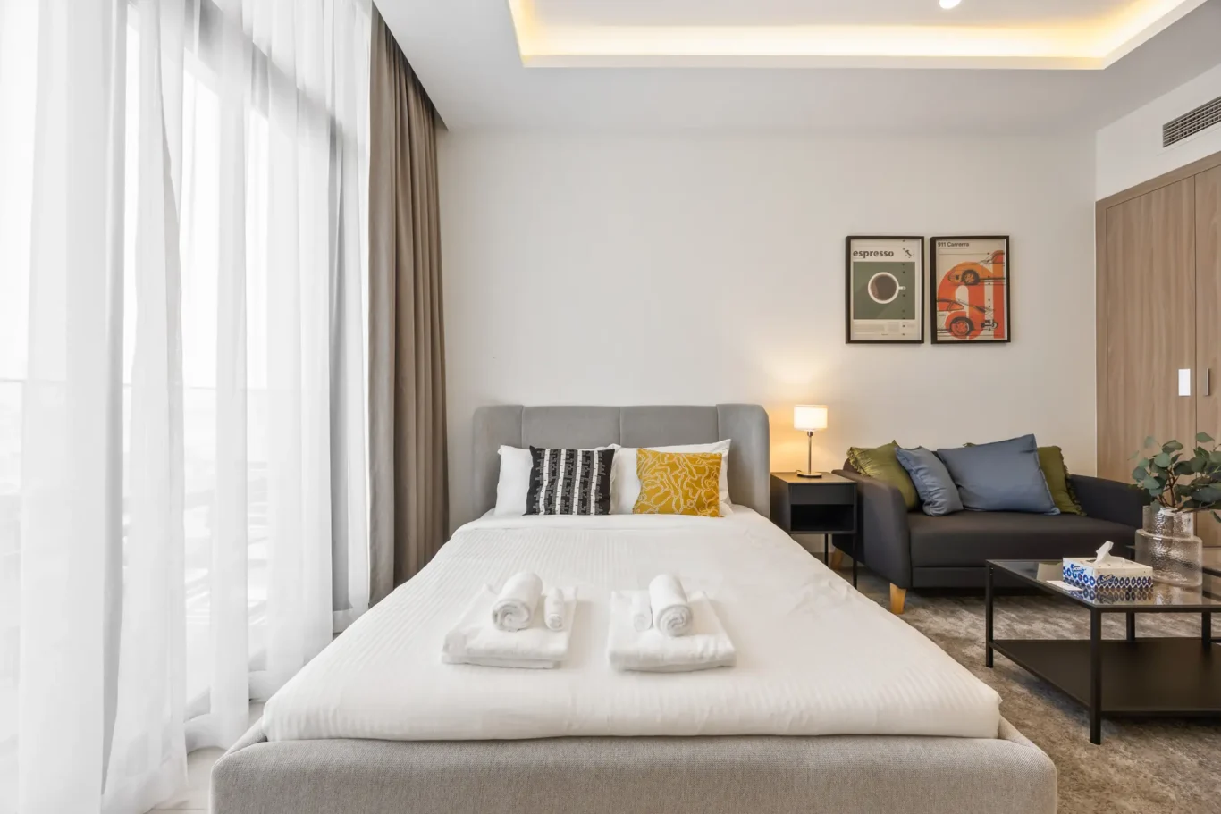 Hotel room with a bed, grey headboard, decorative pillows, sheer curtains, and a small seating area nearby with a sofa and coffee table