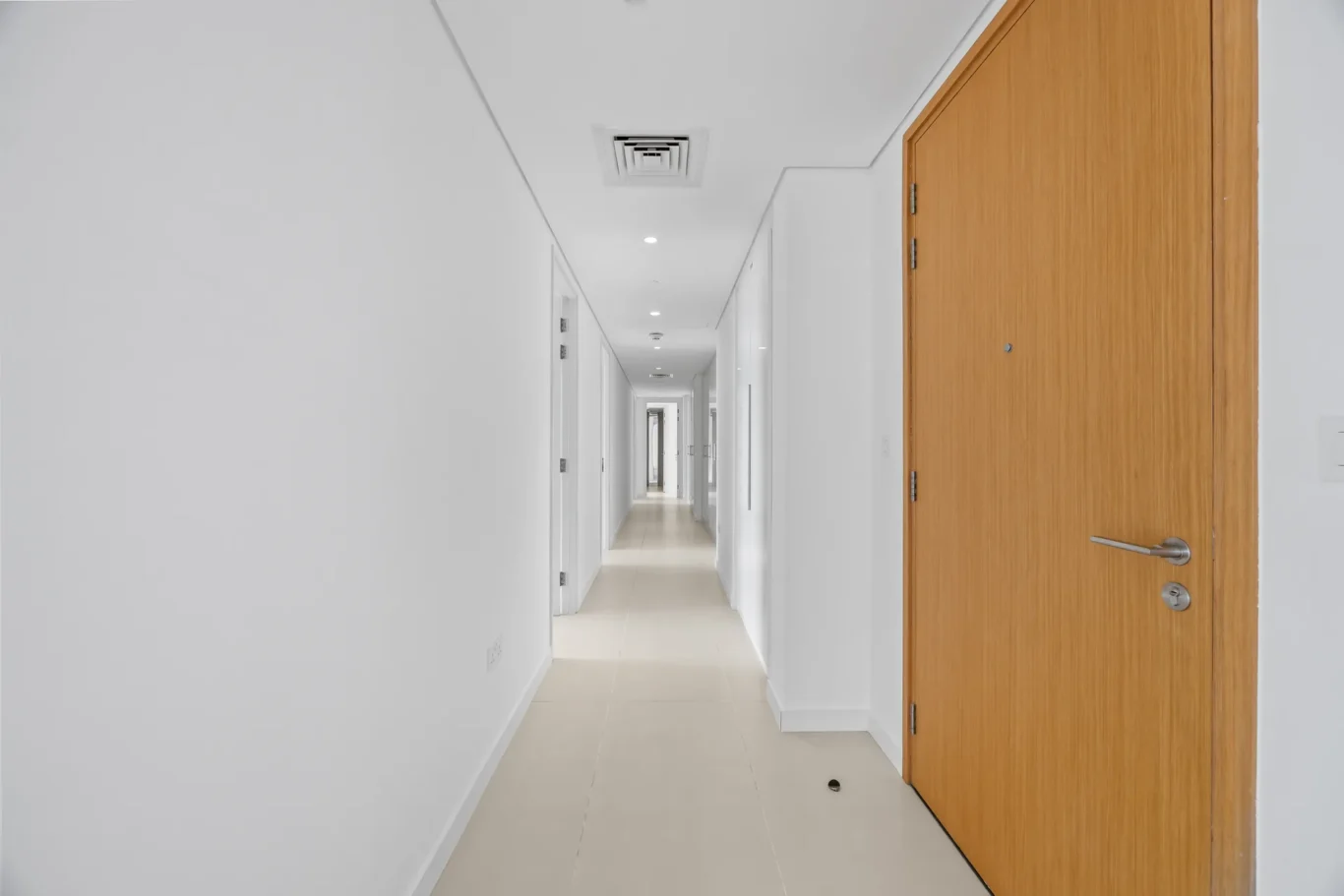Long white hallway with multiple white doors on both sides and a large wooden door at the right, tiled floor.