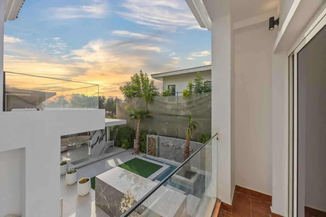 Modern balcony with glass railing, overlooking a landscaped courtyard and palm trees at sunset.
