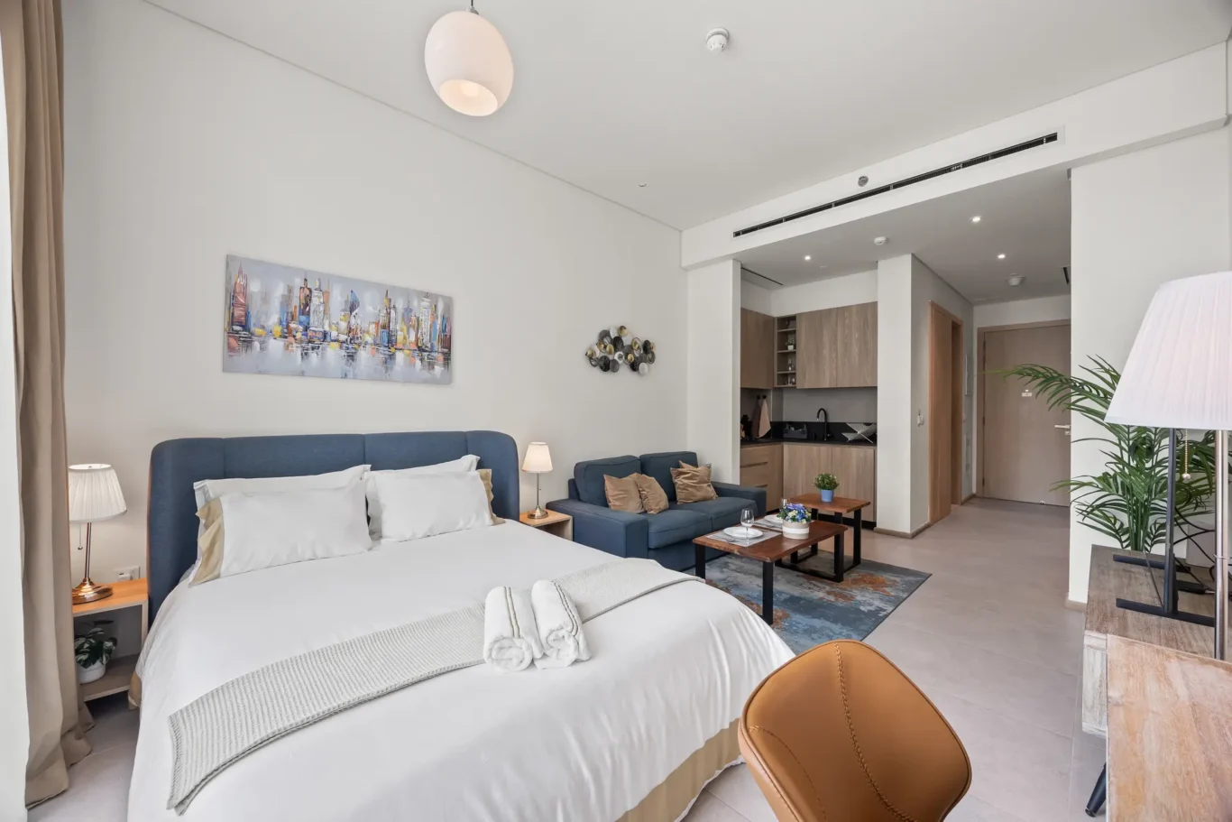 Open-plan hotel suite with a white bed in the foreground, blue headboard, seating area, and a compact kitchenette in the background.