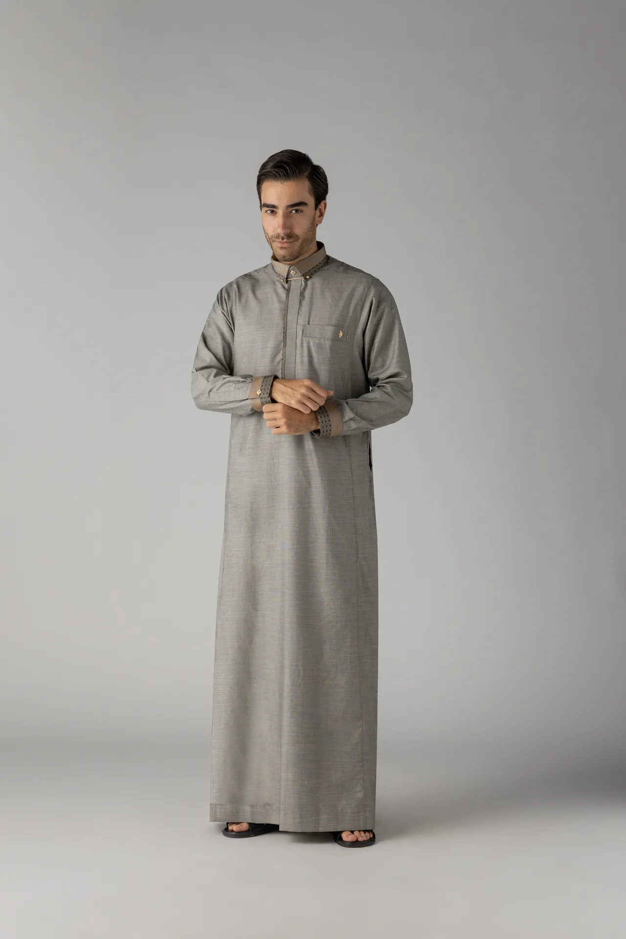 Full-body shot of a man in a long gray thobe/abaya, standing with his hands clasped, wearing black sandals against a gray studio background.