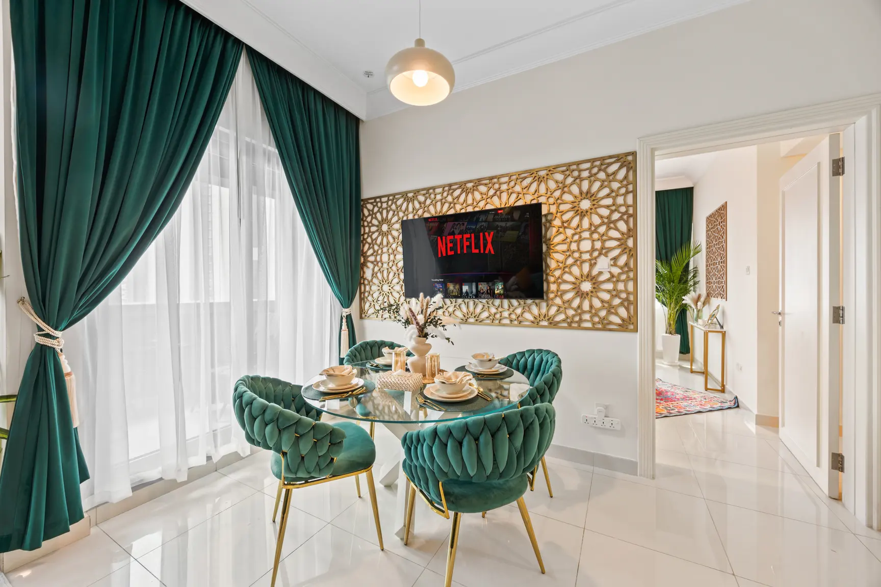 Elegant dining nook: teal velvet chairs around a round glass table, green drapes, and a Netflix screen mounted on a decorative lattice wall.