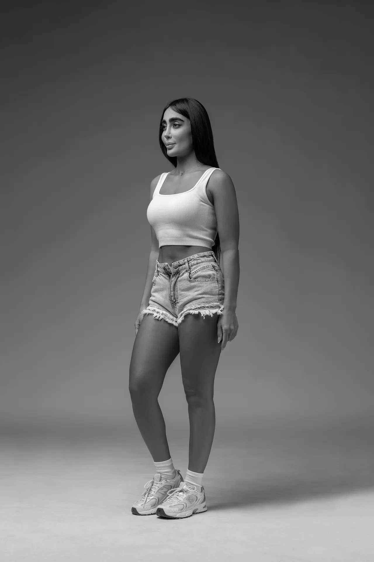 Full-body black-and-white portrait of a woman standing in a white ribbed tank top, frayed denim shorts, and sneakers in a studio setting.