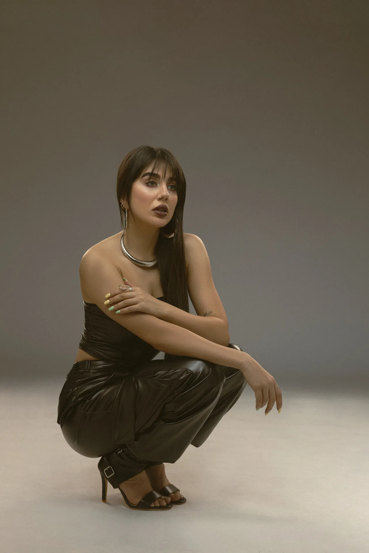 Woman in a black shiny off-shoulder top and pants squatting in a studio, wearing bold makeup and silver jewelry.