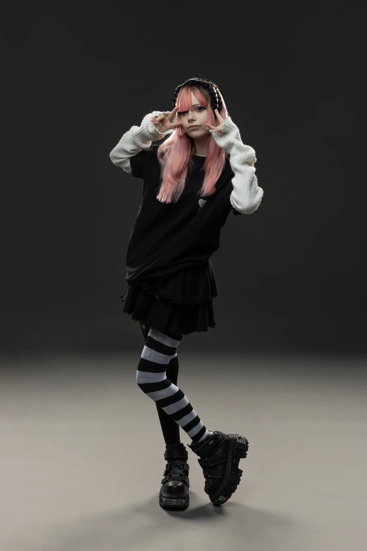 Person with pink hair and black layered outfit posing with peace signs against a dark backdrop, wearing striped tights and chunky boots.