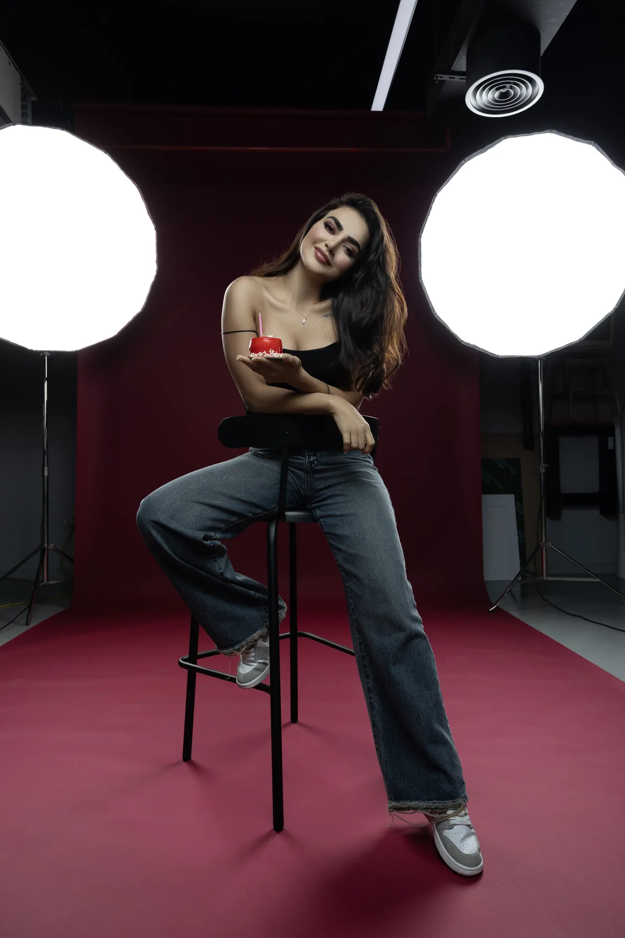 Smiling woman sits on a stool in a photography studio, holding a small red cake with a candle between two bright studio lights.