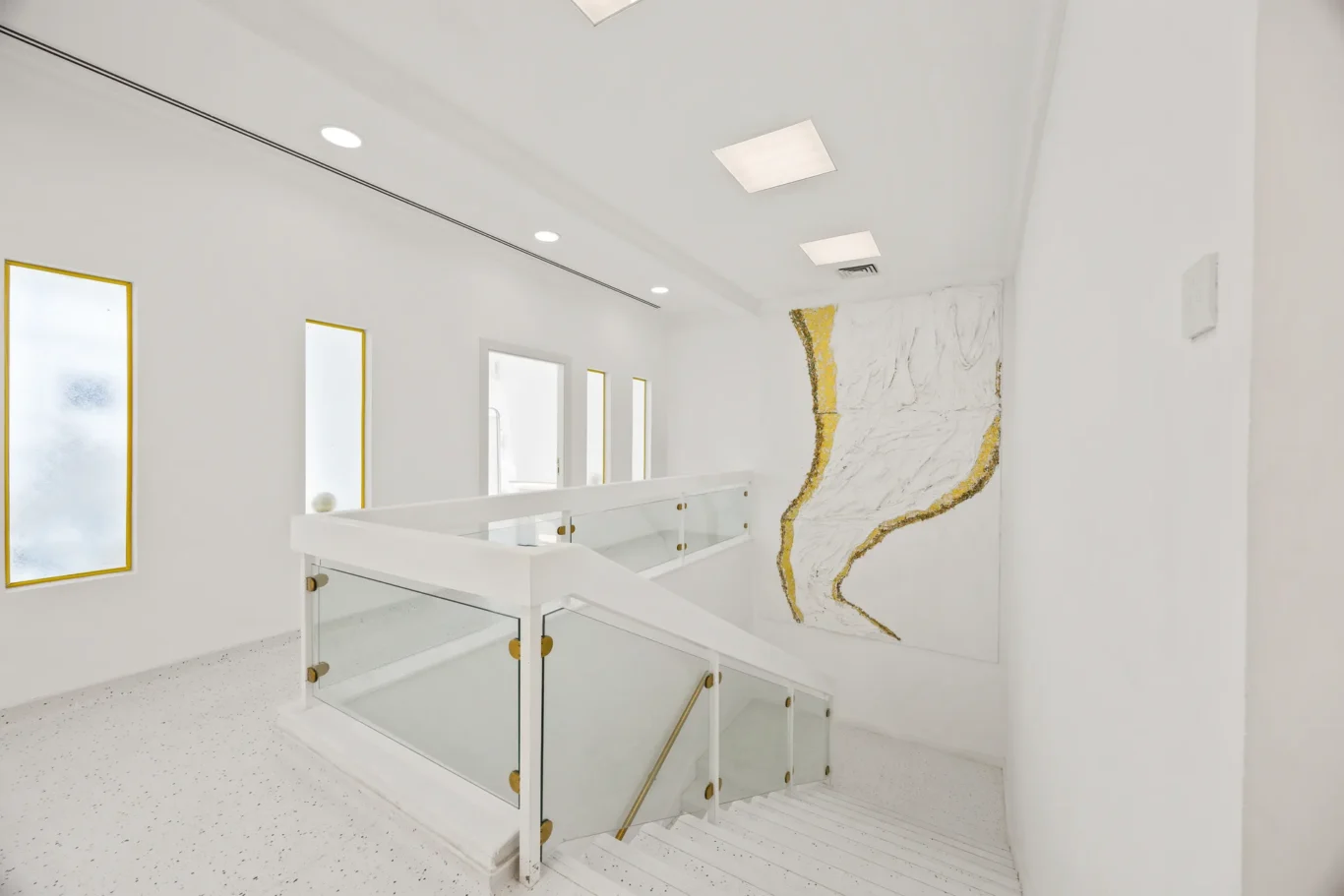 Bright white hallway with glass-railed staircase, frosted windows on the left and a gold-accent abstract mural on the right.
