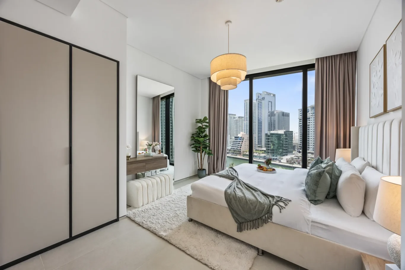 Modern hotel room with a large window overlooking a city skyline, king bed with white linens and gray throw blankets, chandelier overhead, neutral decor.