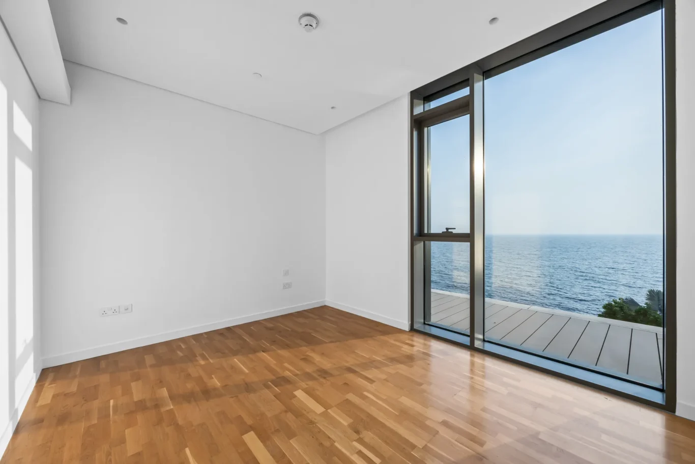 Empty white room with a wooden floor and a large ocean-facing glass door/window viewing the sea.