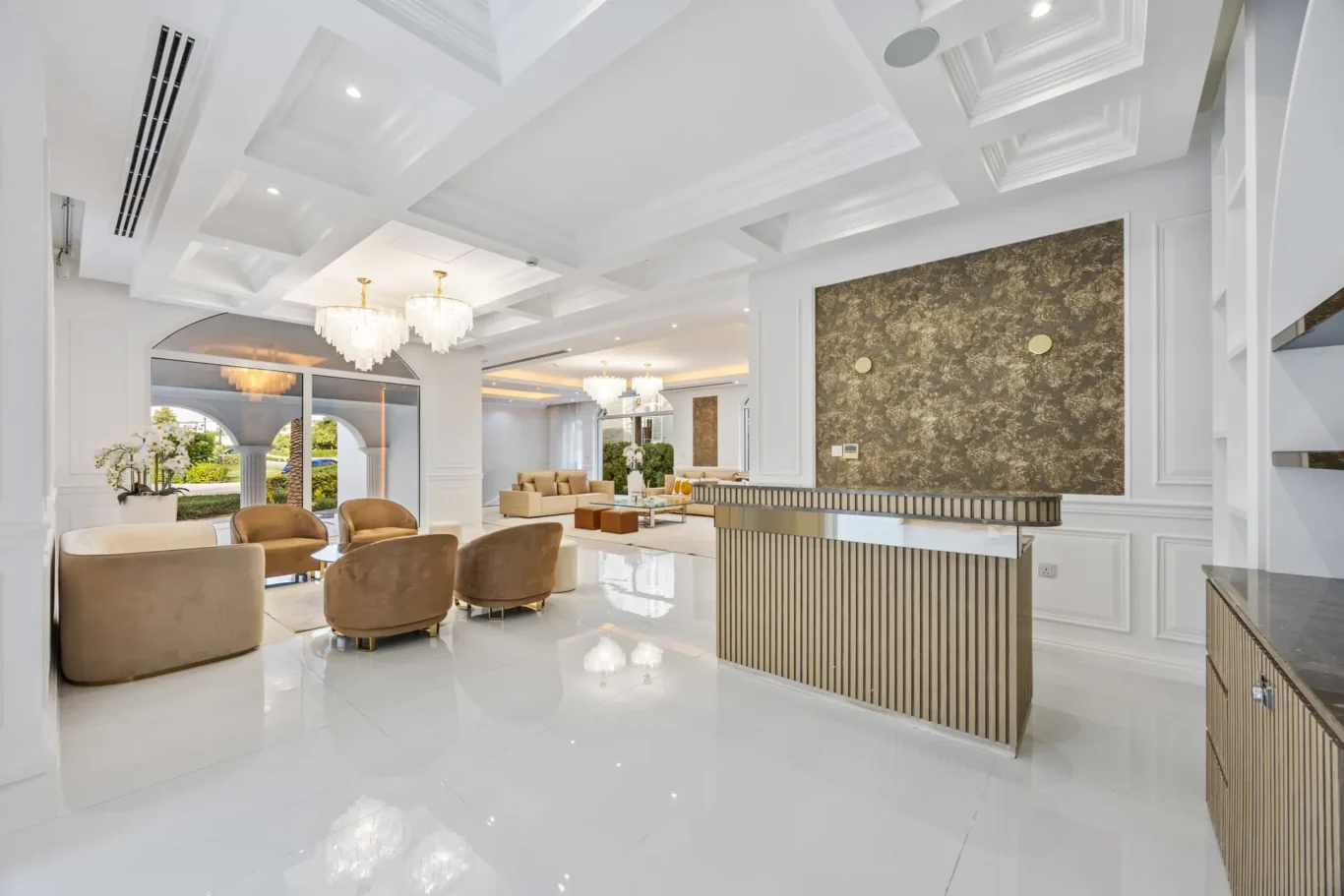 Luxurious white lobby with a coffered ceiling, crystal chandeliers, beige seating, and a striped wood reception desk