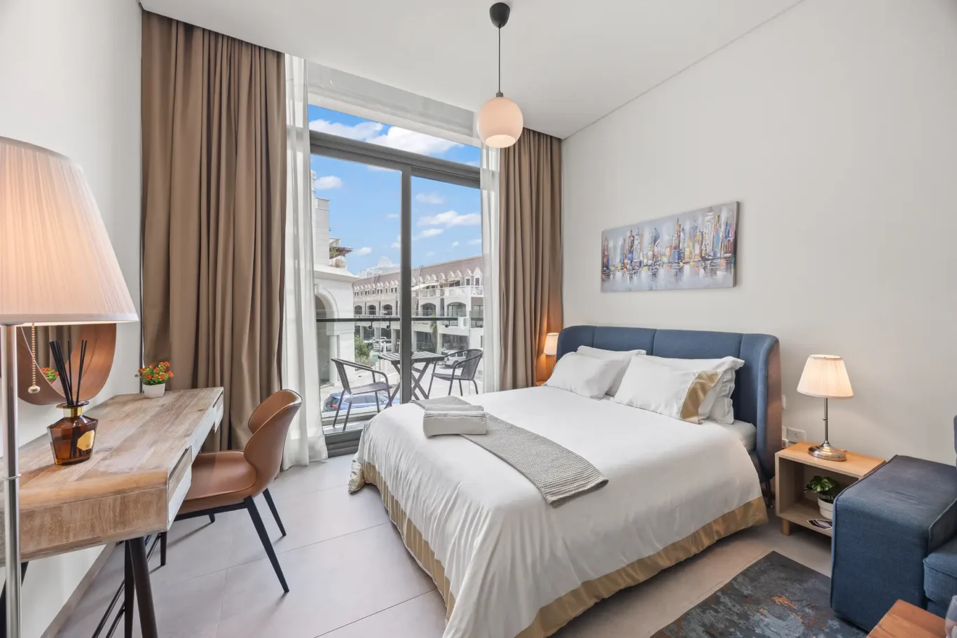 Hotel room with a large window and balcony seating, bed with blue headboard, and a wooden desk on the left.