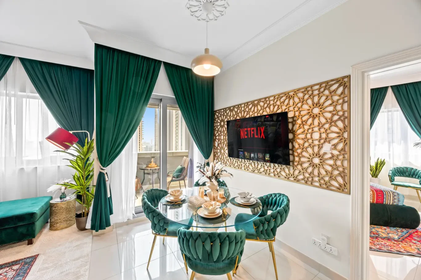 Bright living/dining area with emerald green curtains, teal tufted chairs around a glass dining table, and a decorative wooden geometric screen on the wall.
