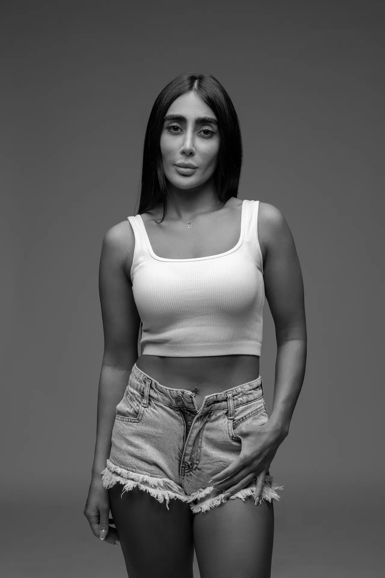 Portrait of a woman in a white crop top and frayed denim shorts, standing against a plain backdrop in black-and-white.