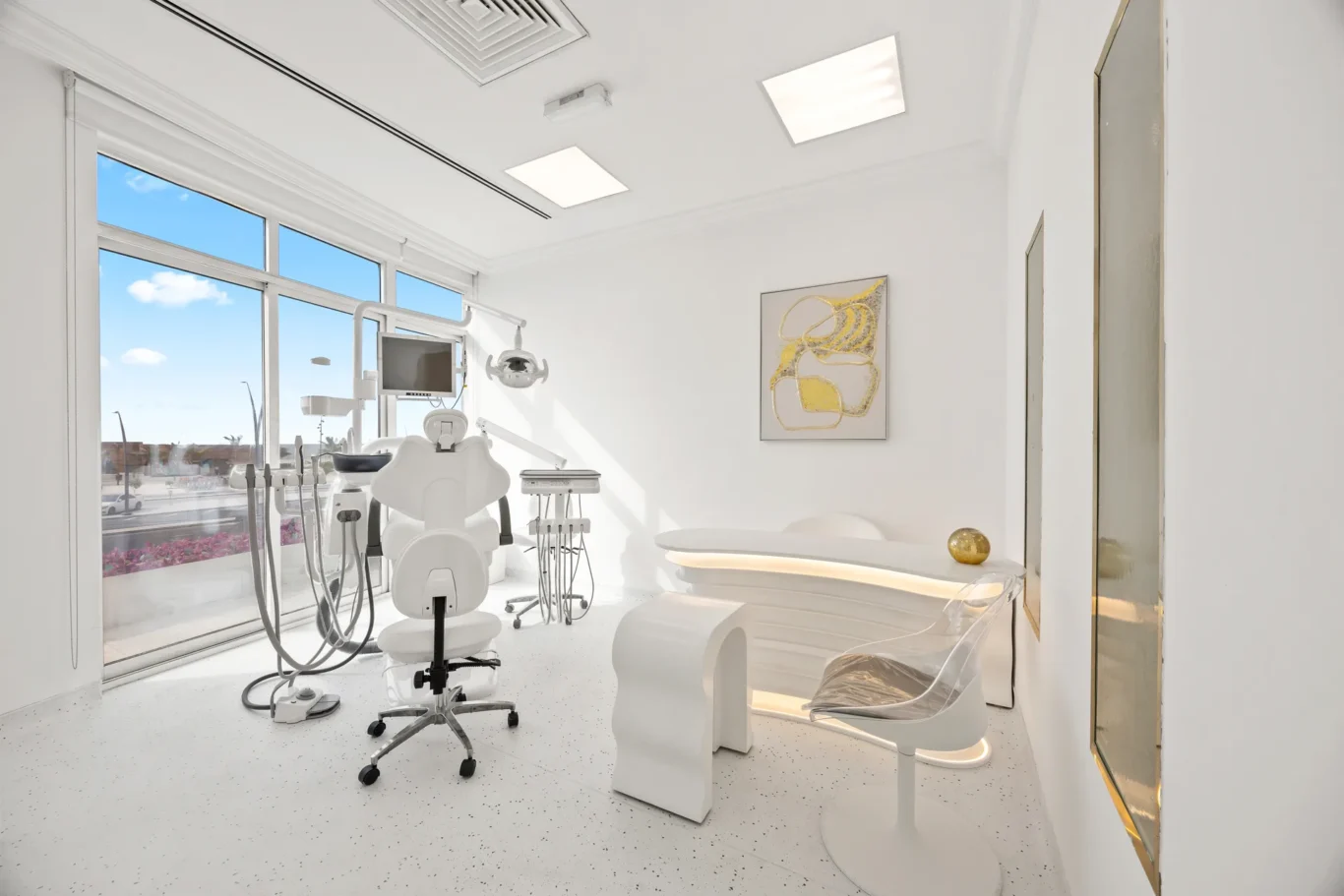 Bright, modern dental treatment room with a white reclining chair, equipment, and large windows letting in daylight.”